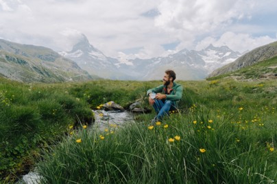 Man camping enjoying his coffee sitting alongside a stream with a beautiful mountain in view