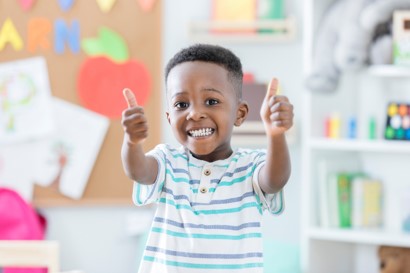 Little boy in a classroom, arms wide open with two thumbs up, smiling in appreciation and joy