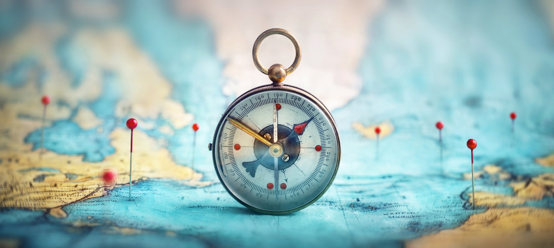 A compass standing vertically on a map which is brightly colored and has various locations highlighted with pushpins